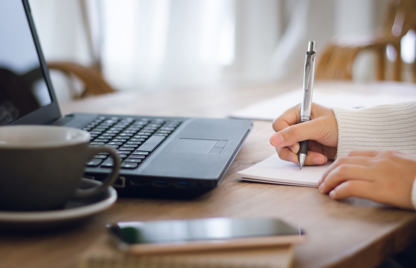 laptop, cup of coffee, person writing on piece of paper
