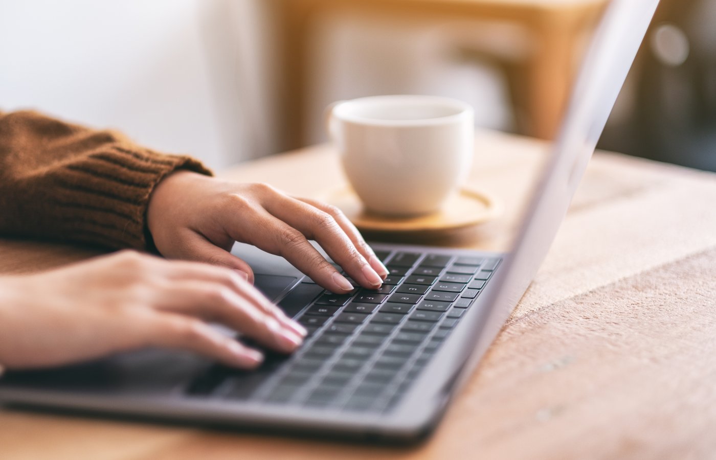 close up of hands typing on laptop with a coffee cup next to it. 
